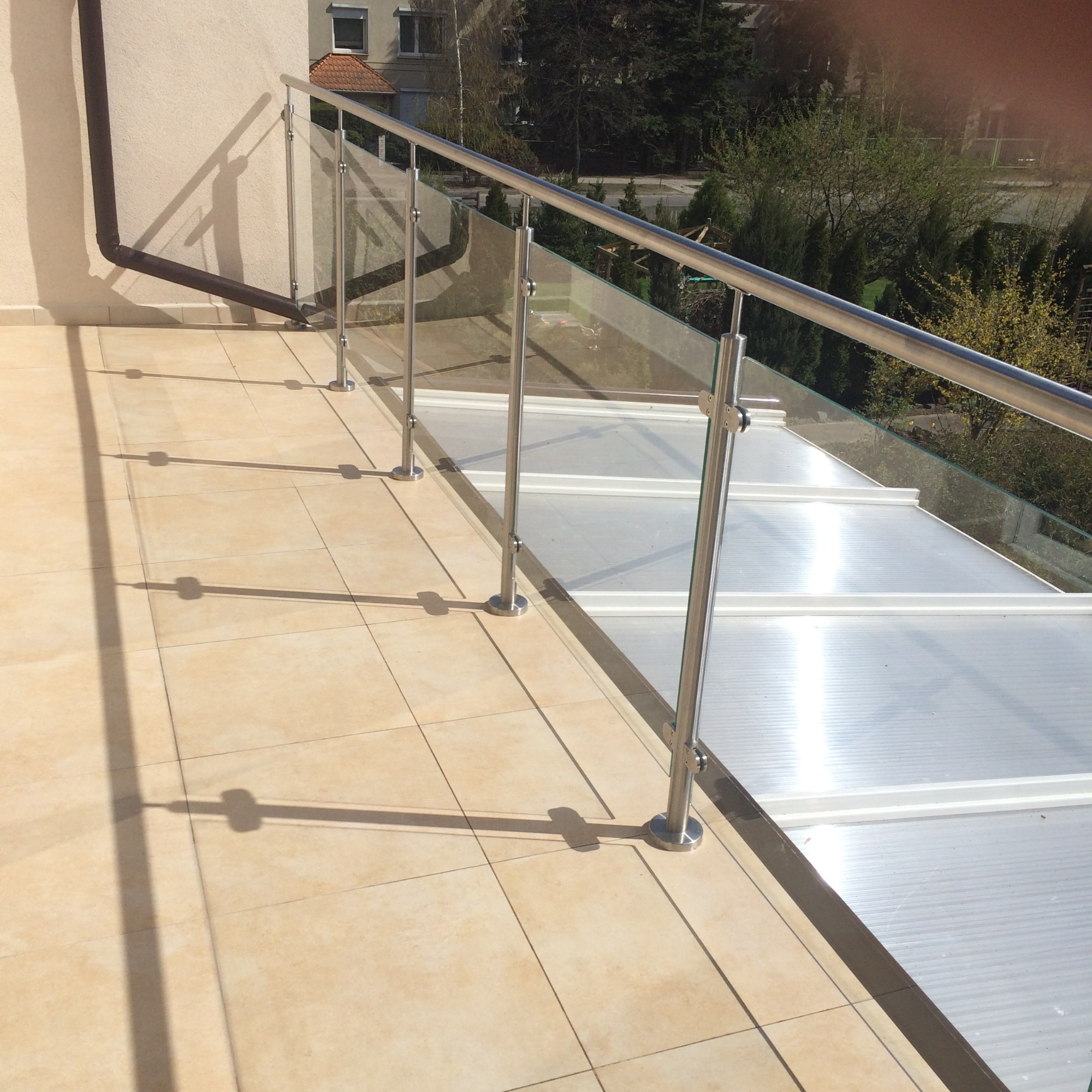 Balkon z nowoczesną, szklaną balustradą ze stali nierdzewnej, oświetlony mocnym słońcem rzucającym wyraźne cienie na jasną, ceramiczną podłogę.