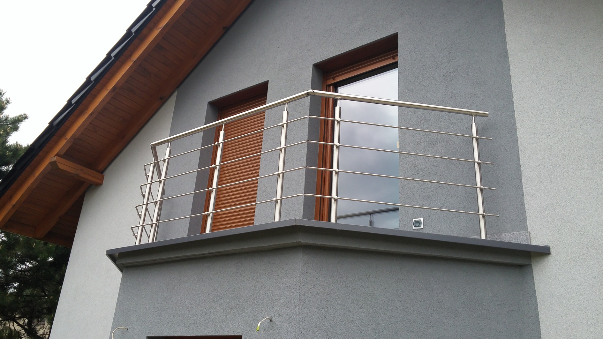 Balkon z balustradą nierdzewną z czterema poprzeczkami, szara elewacja budynku z brązowymi roletami i oknami.