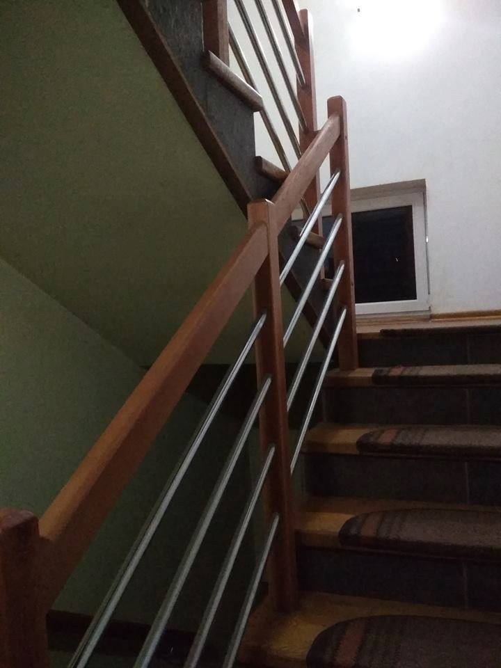 Schody z drewnianą poręczą i metalowymi prętami, widoczne stopnie z nakładkami dywanowymi i okno w tle.