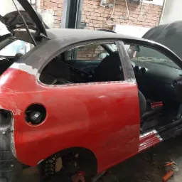 Czerwony samochód w trakcie napraw blacharskich, z otwartą klapą bagażnika i drzwiami, widoczne ślady spawania i szlifowania, uszkodzony tylny błotnik, narzędzia leżące na podłodze w warsztacie.