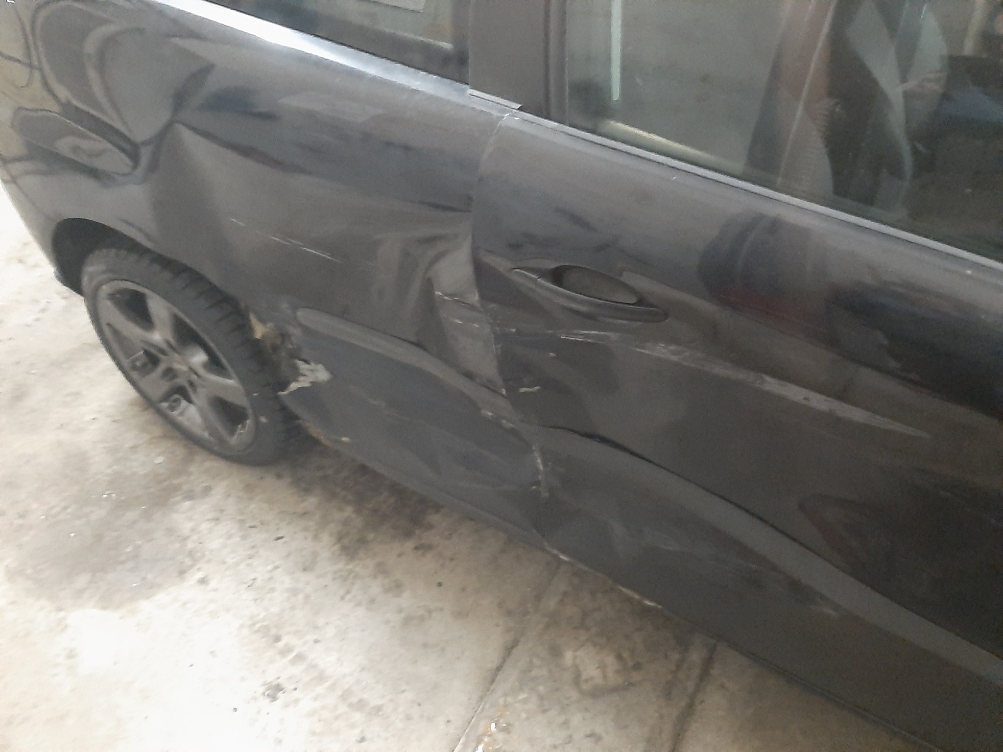 Uszkodzone drzwi i błotnik ciemnego samochodu, widoczne wgniecenia i zarysowania lakieru, zarysowania na oponie.