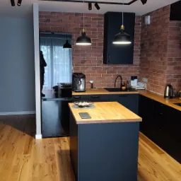 Nowoczesna kuchnia z ceglaną ścianą, drewnianym blatem wyspy i czarnymi szafkami, oświetlona lampami wiszącymi i reflektorami na szynoprzewodzie.