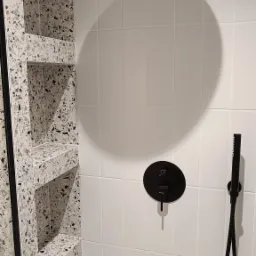 Białe, kwadratowe płytki na ścianie kabiny prysznicowej z czarną, okrągłą baterią i słuchawką, obok wnęka w ścianie wykończona płytkami lastryko.