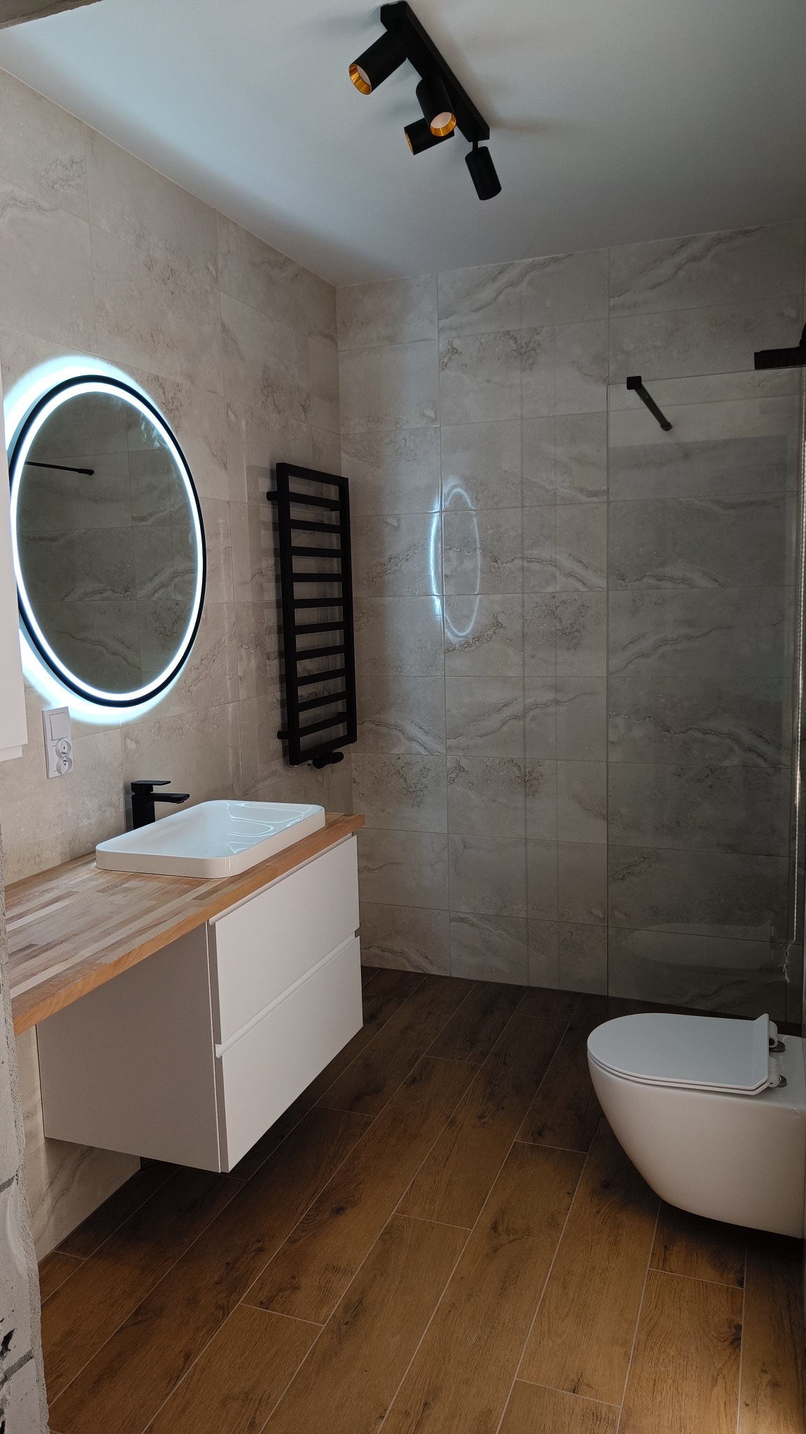 Nowoczesna łazienka z drewnopodobną podłogą, umywalką nablatową z okrągłym lustrem LED, szklaną kabiną prysznicową i grzejnikiem drabinkowym w kolorze czarnym.