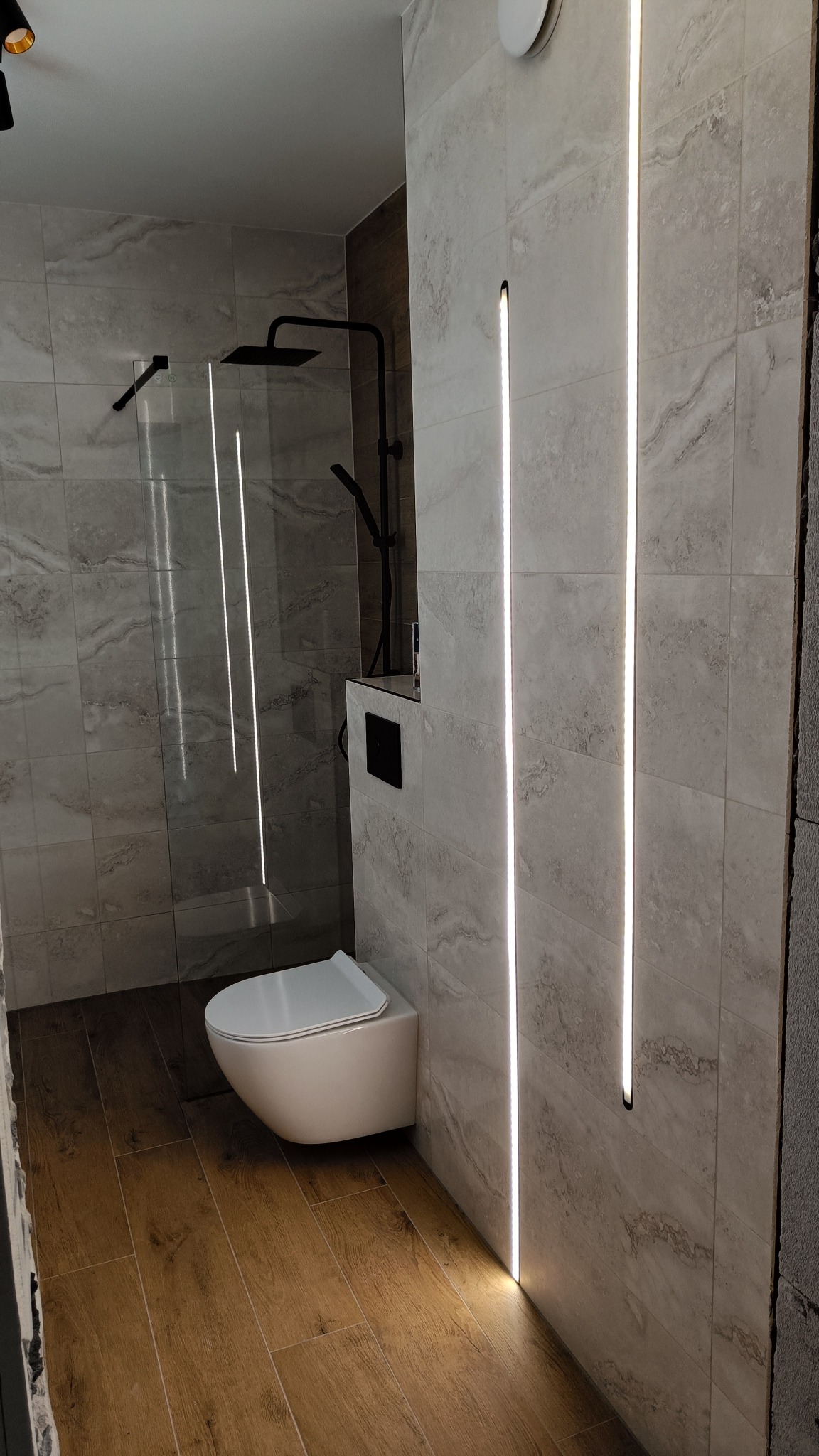 Nowoczesna łazienka z podświetlanymi panelami ściennymi, wiszącą toaletą i kabiną prysznicową typu walk-in z czarną armaturą.