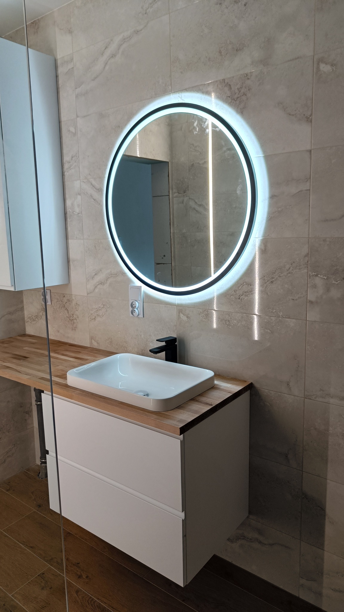 Nowoczesna łazienka z okrągłym lustrem LED, białą umywalką nablatową i drewnianym blatem, płytki ścienne w odcieniach beżu i szarości z marmurkowym wzorem.