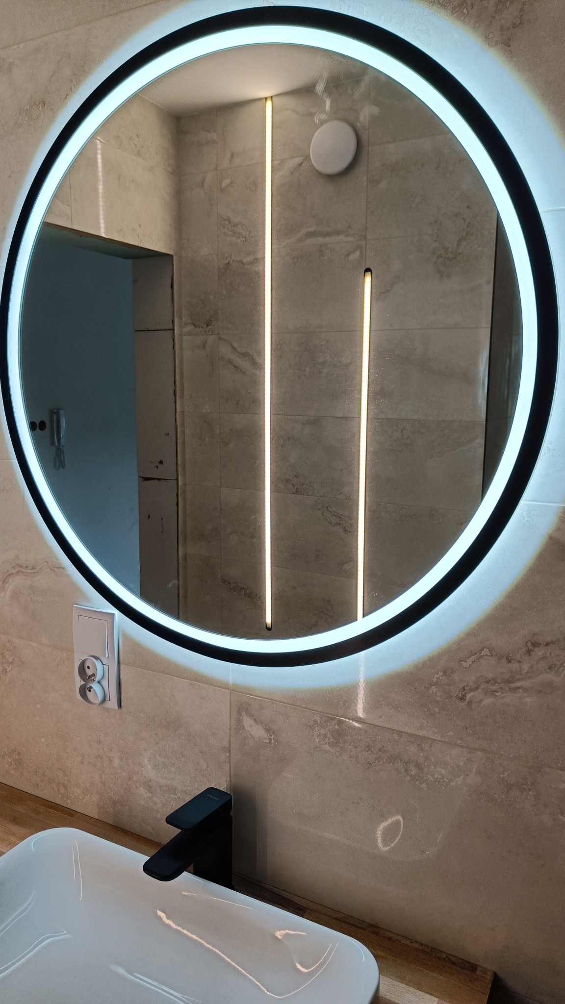 Okrągłe lustro LED z podświetleniem na płytkach imitujących marmur, widoczna biała umywalka i czarna bateria, w tle fragment otwartych drzwi.