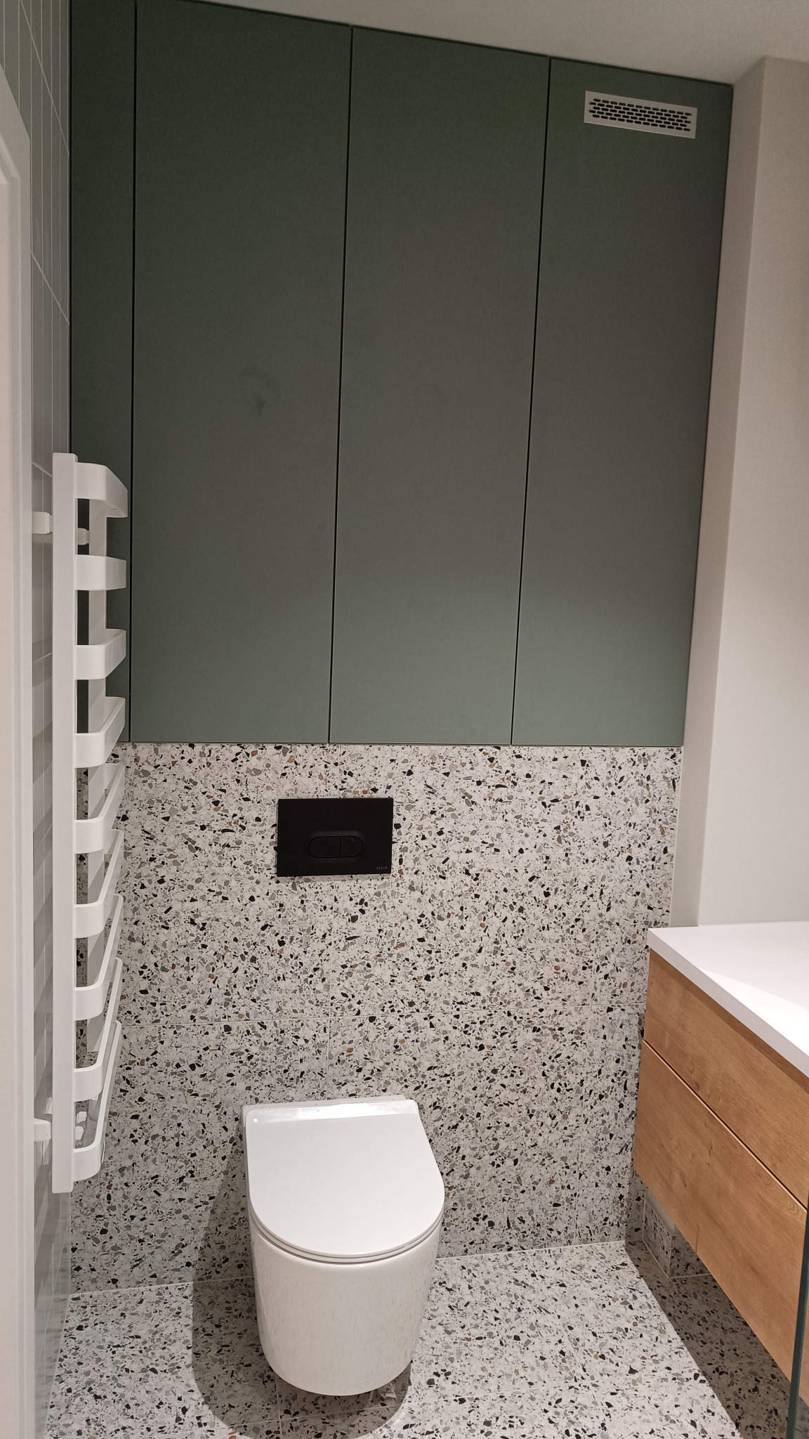 Nowoczesna łazienka z toaletą wiszącą, płytkami lastryko na ścianach i podłodze, zabudową meblową w kolorze oliwkowym i drewnianą szafką pod umywalką.