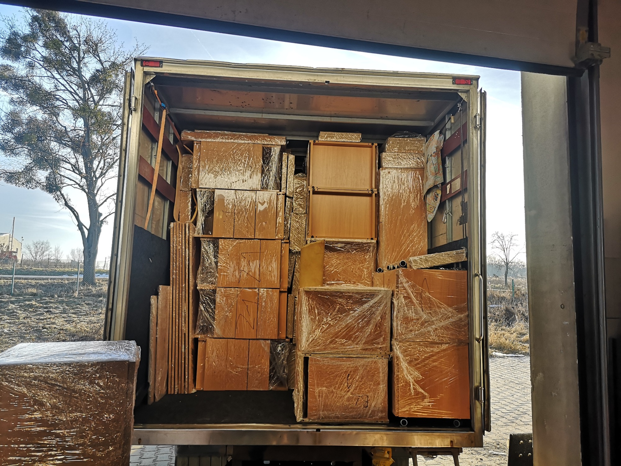 Wnętrze ciężarówki wypełnione meblami zabezpieczonymi folią stretch, widoczne elementy szafek i płyty meblowe, załadunek w słoneczny dzień, drzewo bez liści w tle.