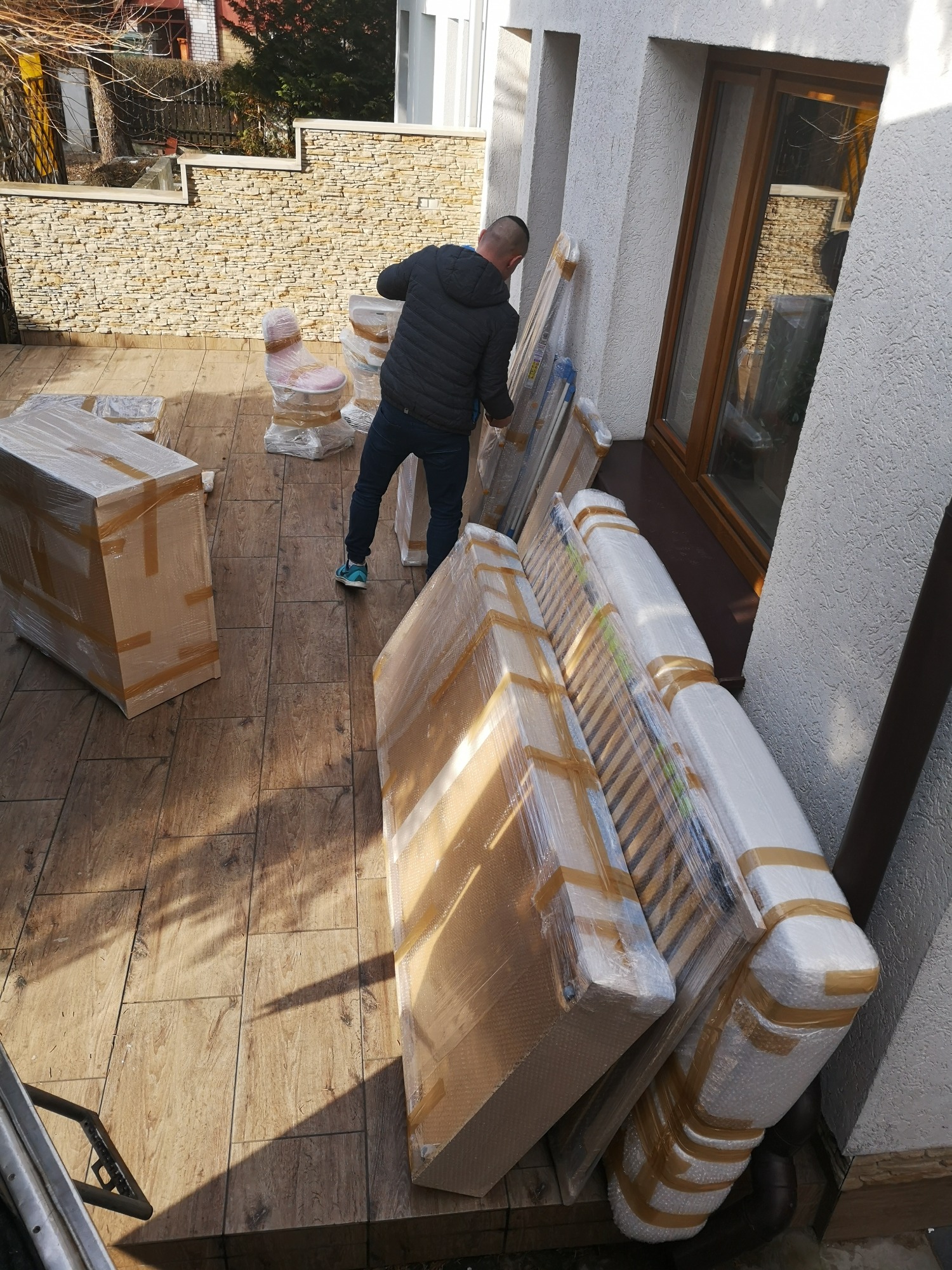 Mężczyzna w ciemnej kurtce i dżinsach przenosi meble zabezpieczone folią i taśmą na zewnątrz budynku z kamienną elewacją. Widoczne krzesła, materace i skrzynie.