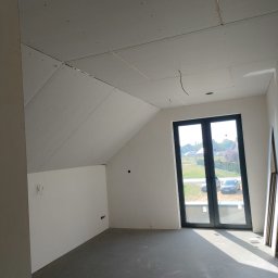 Usługi remontowo budowlane - Poddasze z oknem, ściany częściowo wykończone płytami gipsowo-kartonowymi, widoczne przewody i puszki elektryczne, surowa podłoga. Etap prac wykończeniowych.