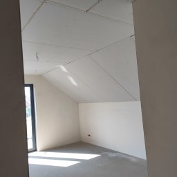 Usługi remontowo budowlane - Pokój na poddaszu z zamontowanymi płytami karton-gips na skośnym suficie, widok z perspektywy wejścia, surowy stan wykończenia, okno z czarną ramą.