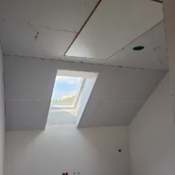 Montaż płyt gipsowo-kartonowych na poddaszu z oknem dachowym. Widoczne łączenia płyt i otwór rewizyjny w suficie. Świeże, białe ściany w pomieszczeniu.