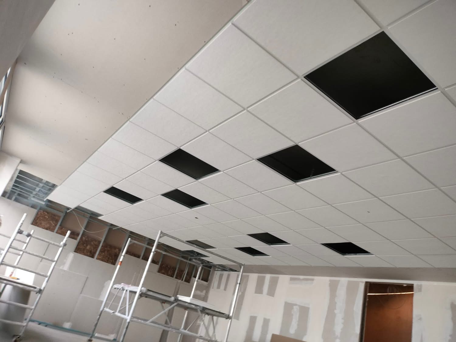 Montaż sufitu podwieszanego z płyt w jasnym pomieszczeniu. Widoczne rusztowania i otwory w płytach. Prace wykończeniowe w trakcie realizacji.