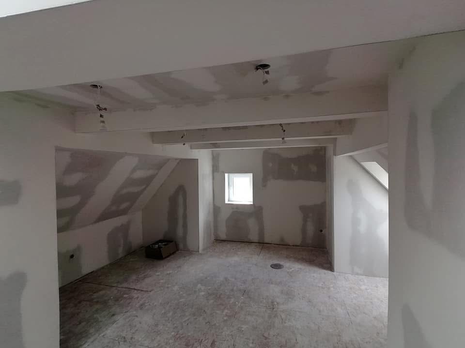 Pokój na poddaszu w trakcie prac wykończeniowych, ściany pokryte gładzią, widoczne elementy konstrukcyjne sufitu i małe okno.