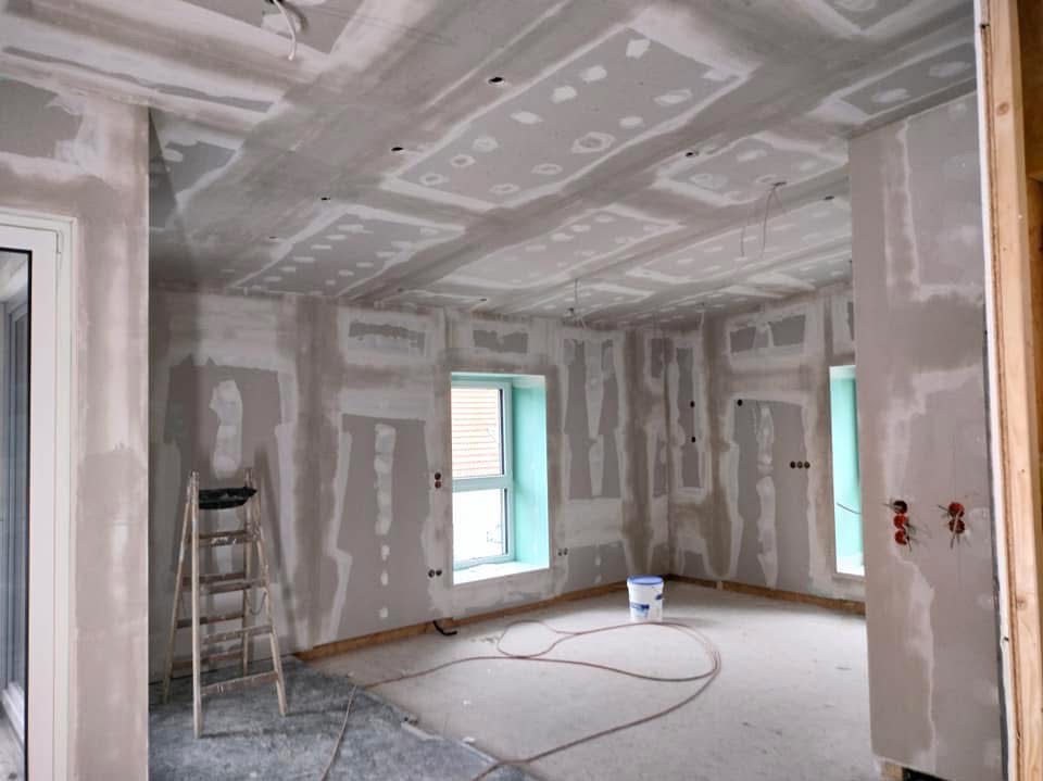 Pokój w trakcie prac wykończeniowych: ściany i sufit pokryte gładzią, widoczne łączenia płyt karton-gips, drabina, kabel na podłodze, okno z widokiem na zewnątrz.