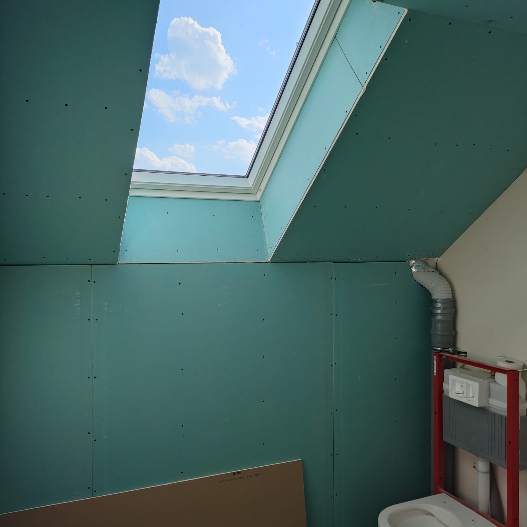 Montaż zielonych płyt karton-gips w łazience na poddaszu ze świetlikiem. Widoczna instalacja toalety podtynkowej i fragment niebieskiego nieba.