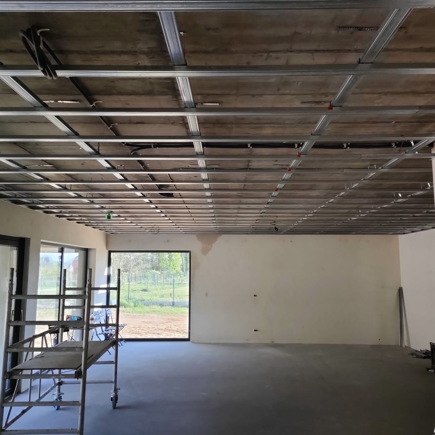 Montaż sufitu podwieszanego z metalową konstrukcją w Otyniu. Widoczne kable i drabina. Ściany przygotowane do dalszych prac wykończeniowych.