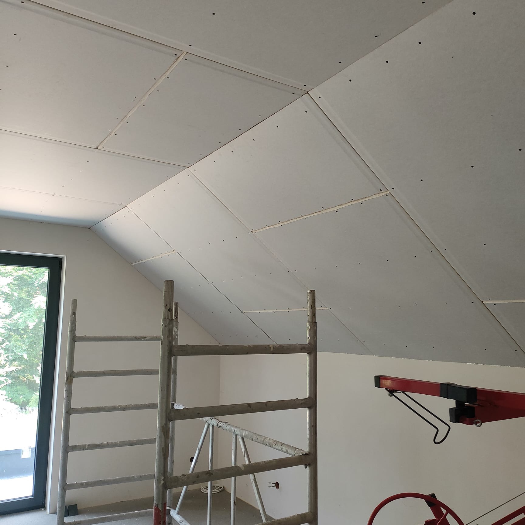 Montaż płyt na skośnym poddaszu z oknem. Widoczne rusztowanie i podnośnik do płyt. Częściowo zakończony montaż płyt.