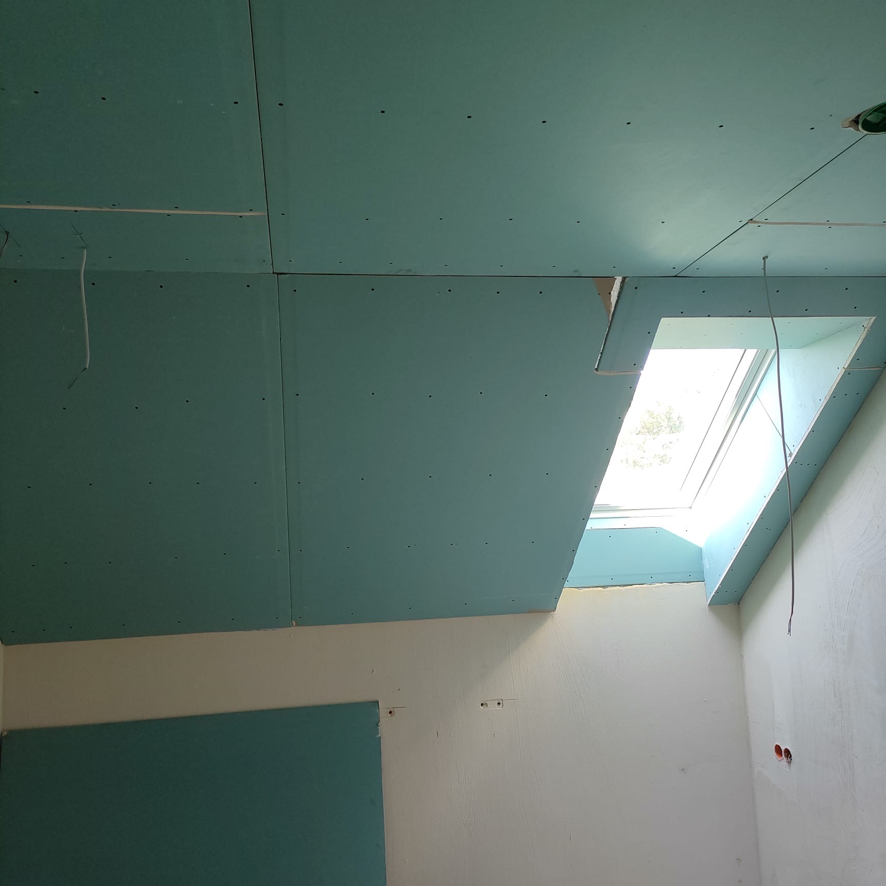 Montaż płyt G-K wokół okna dachowego. Widoczne fragmenty ścian i sufitu z zamontowanymi płytami, wystające przewody. Etap prac wykończeniowych.