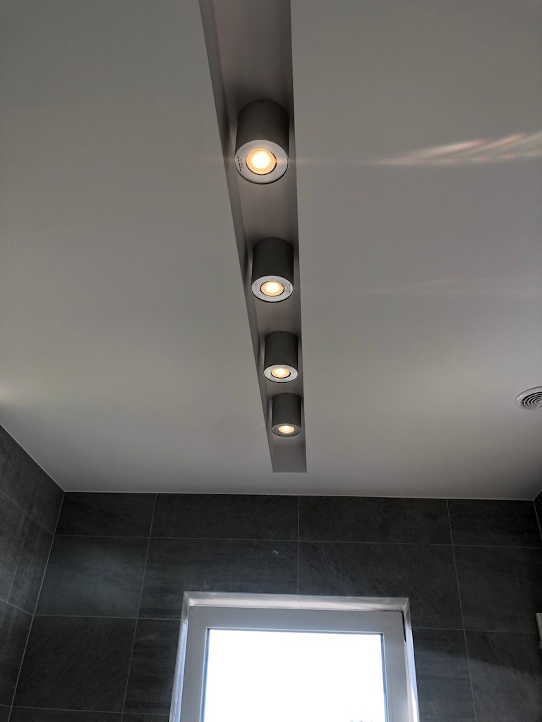 Szara listwa z pięcioma punktowymi lampami LED w suficie, szare płytki na ścianie, biała rama okienna