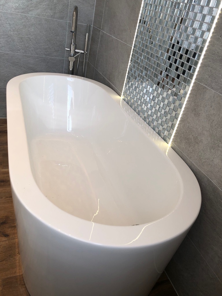 Wolnostojąca biała wanna z chromowaną armaturą na tle ściany wyłożonej szarą płytką i mozaiką lustrzaną z podświetleniem LED.