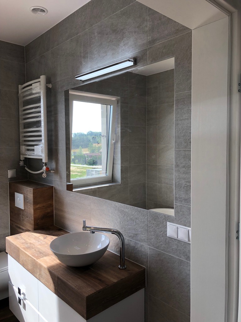 Nowoczesna łazienka z szarymi płytkami, drewnianym blatem pod umywalką i grzejnikiem drabinkowym w folii ochronnej, widok na okno z zielenią.