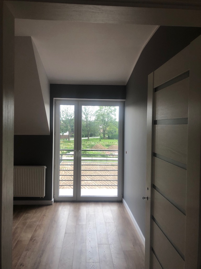 Jasne wnętrze po remoncie: drewnopodobna podłoga, białe drzwi balkonowe z widokiem na zieleń, szare ściany i biały grzejnik, widoczne przez framugę drzwi.