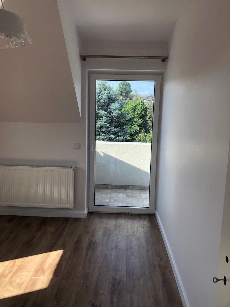 Pokój na poddaszu z drewnianą podłogą, białymi ścianami, grzejnikiem i balkonem z widokiem na drzewa.