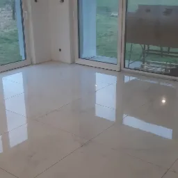 Błyszcząca, biała podłoga z płytek w nowym, pustym pomieszczeniu z dużymi oknami, przez które widać trawnik.