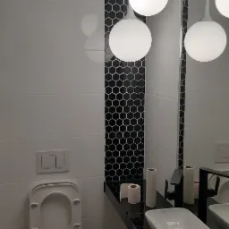 Nowoczesna łazienka z białymi płytkami, czarnym pasem dekoracyjnym w heksagony i wiszącymi lampami kulami, widoczna toaleta podwieszana z otwartą klapą i umywalka nablatowa z czarną armaturą.