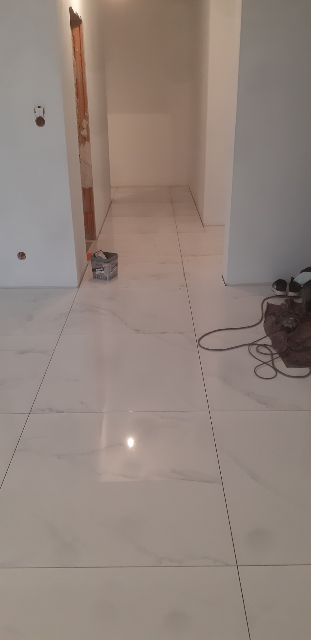 Białe płytki podłogowe imitujące marmur, świeżo ułożone w przejściu między pomieszczeniami, widoczny pojemnik po kleju i narzędzia pracy.