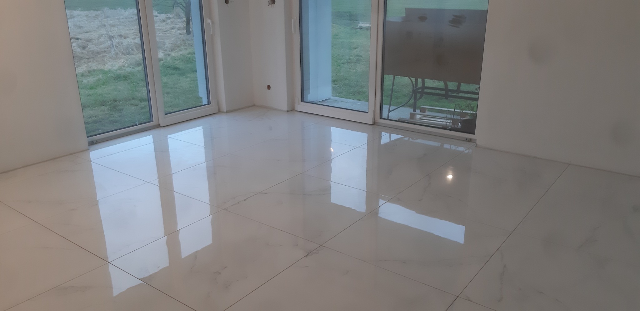 Błyszcząca, biała podłoga z płytek w nowym, pustym pomieszczeniu z dużymi oknami, przez które widać trawnik.