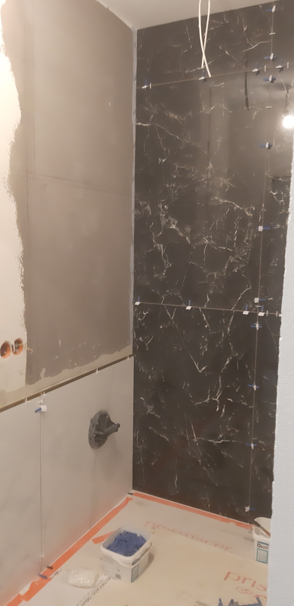 Fragment łazienki w trakcie prac wykończeniowych: czarne płytki imitujące marmur na ścianie, narzędzia glazurnicze, obudowa wanny z zamontowaną baterią, przygotowana do dalszych prac.