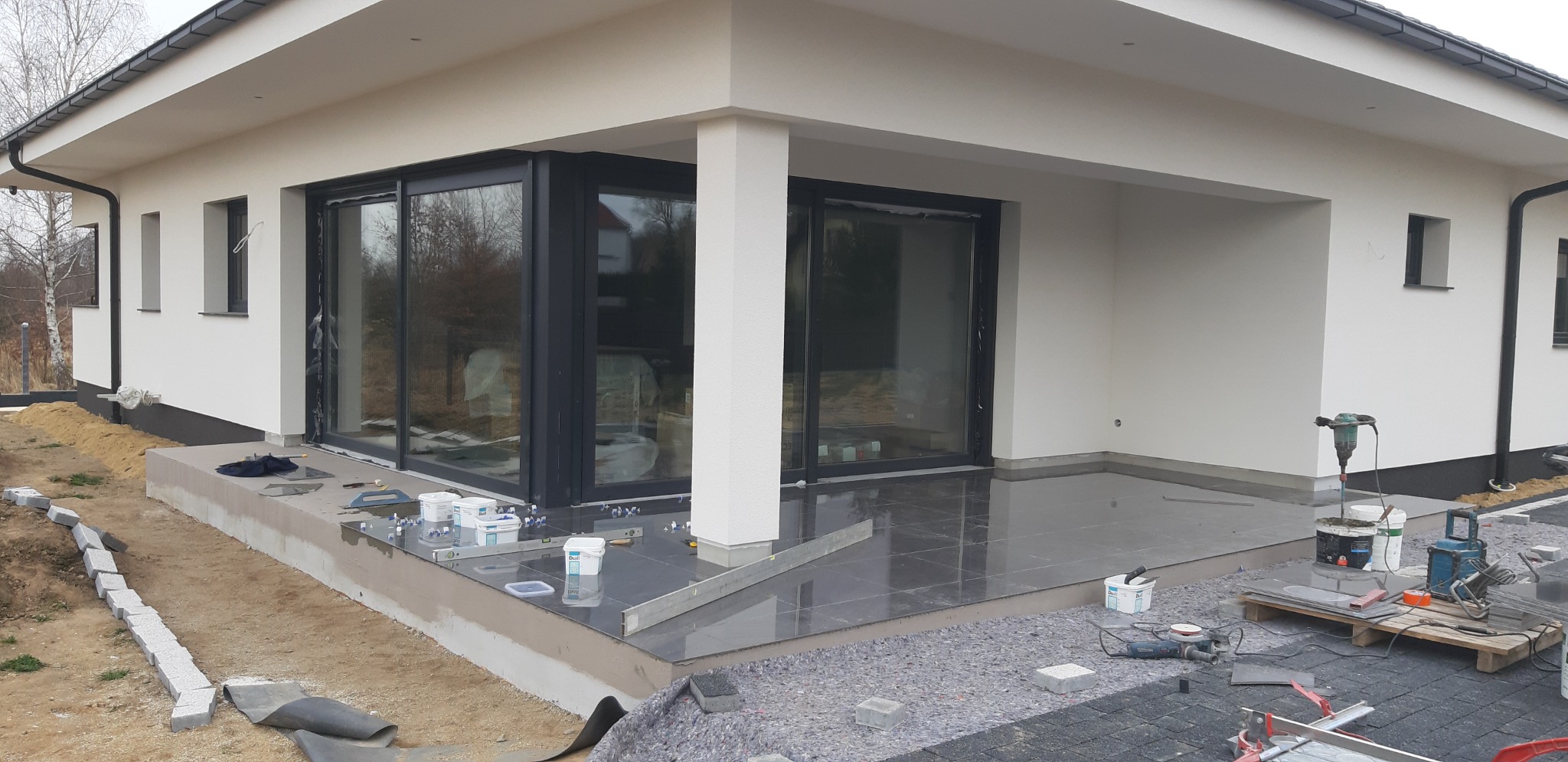 Układanie ciemnych płytek na tarasie domu jednorodzinnego, widoczne narzędzia, poziomica, zaprawa i kostka brukowa w tle.