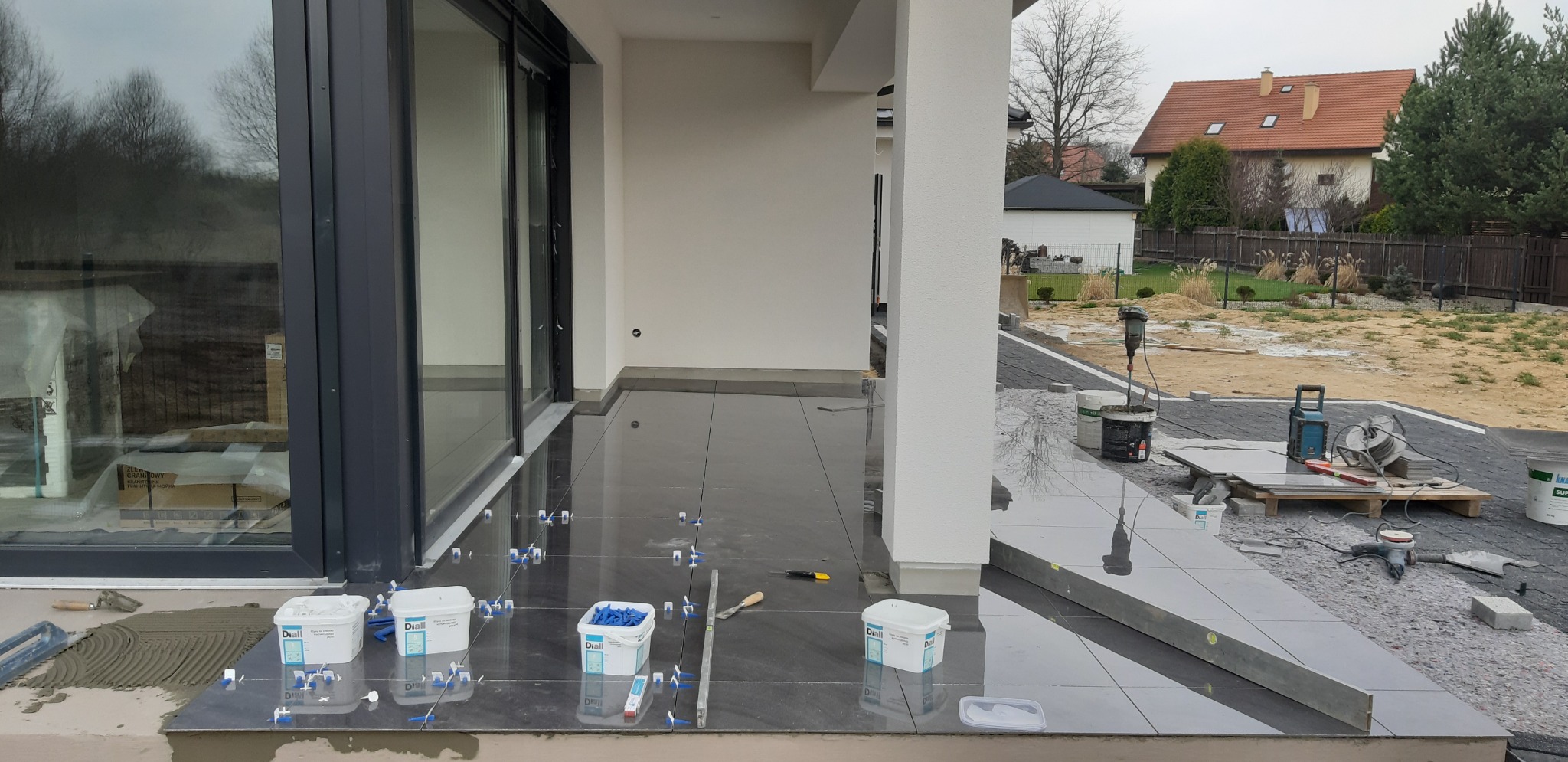 Układanie ciemnych, błyszczących płytek na tarasie zewnętrznym domu jednorodzinnego, widoczne narzędzia i materiały glazurnicze, system poziomowania płytek.