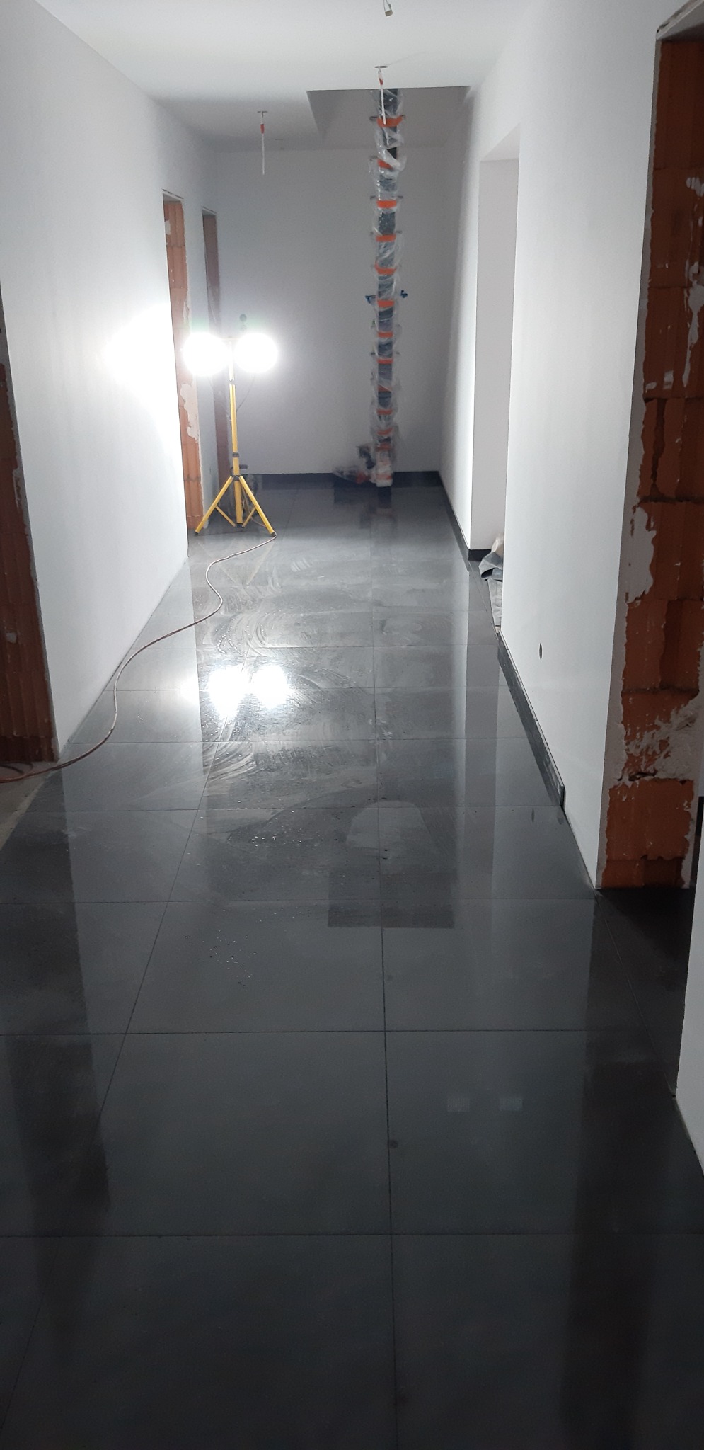 Położona, lśniąca, grafitowa podłoga z płytek w korytarzu, widoczne kable w peszlach pod sufitem, lampa budowlana na trójnogu, surowe ściany w trakcie wykończenia.