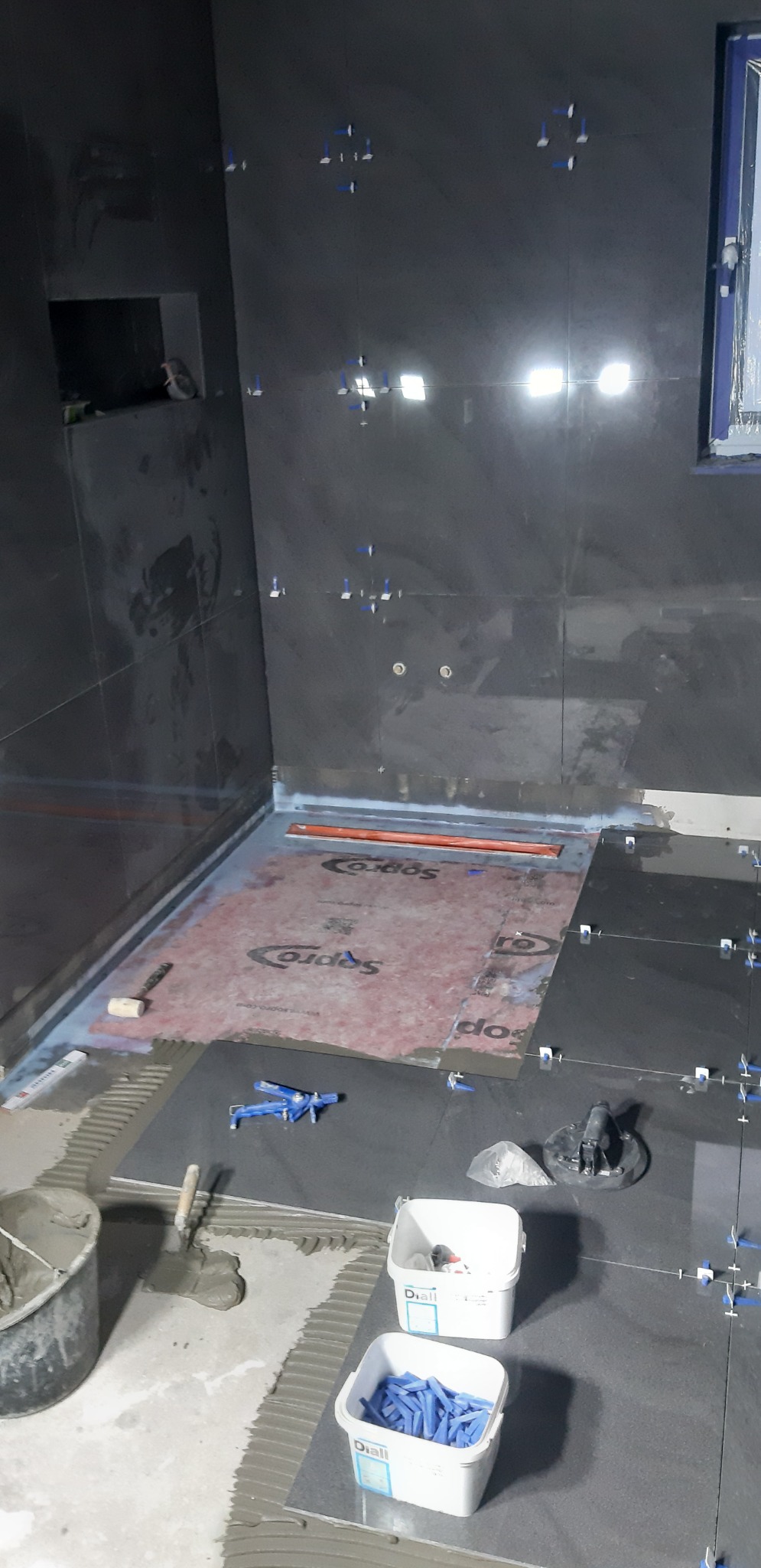 Montaż ciemnych płytek w łazience, widoczny odpływ liniowy, hydroizolacja, narzędzia glazurnicze, krzyżyki dystansowe i wiaderka z zaprawą.