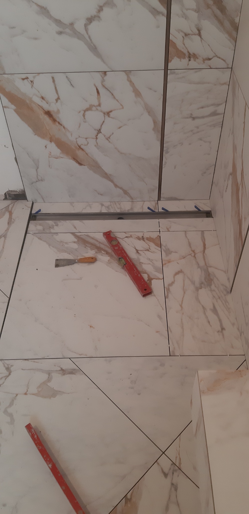 Układanie płytek imitujących marmur w łazience, widoczny odpływ liniowy, poziomica i szpachelka.