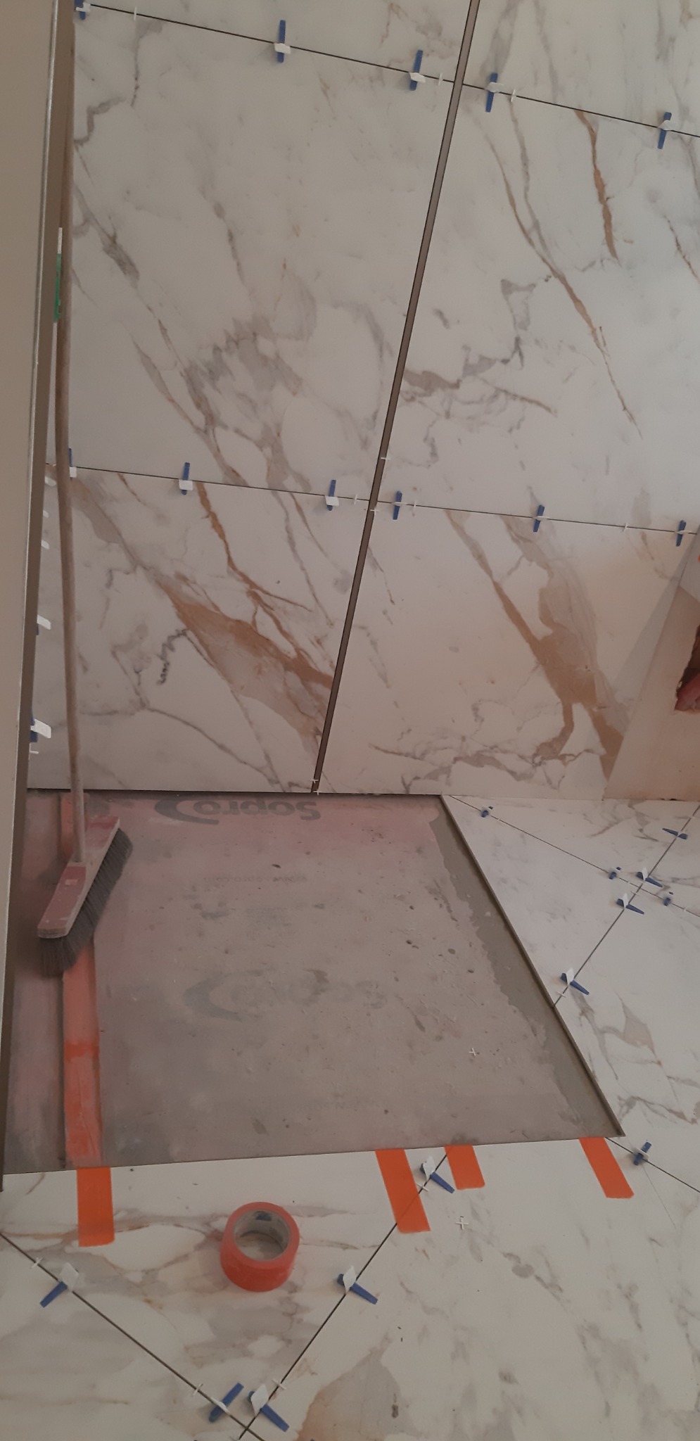Układanie płytek imitujących marmur na ścianie i podłodze łazienki, widoczne krzyżyki dystansowe, taśma malarska i szczotka do zamiatania