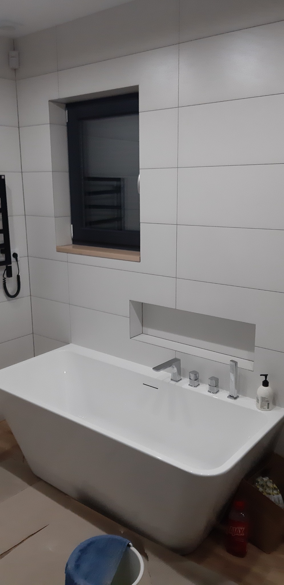 Nowoczesna, wolnostojąca wanna z chromowaną armaturą i wnęką w ścianie wyłożonej białymi, prostokątnymi płytkami. Widoczne okno z czarną ramą i grzejnik drabinkowy. Na podłodze widoczne ślady po...