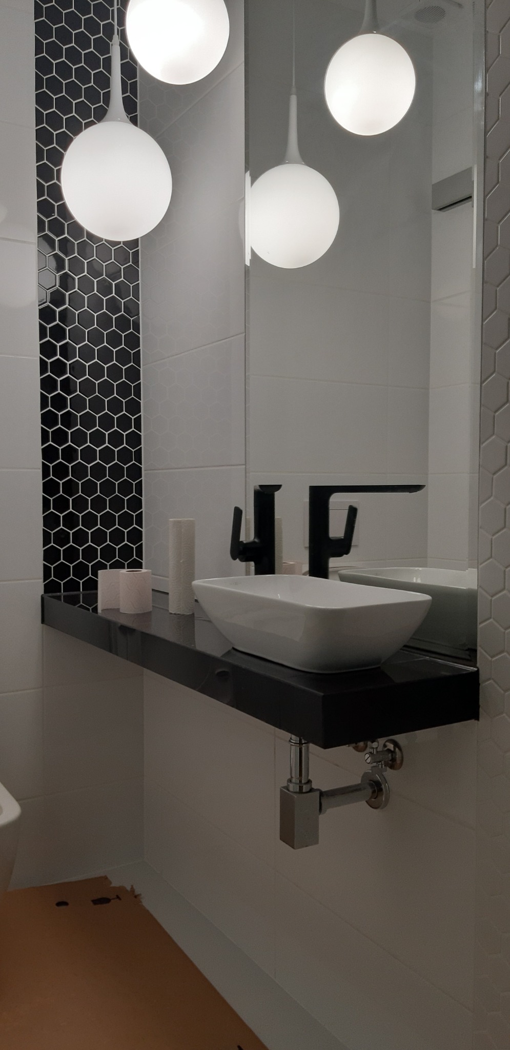 Nowoczesna łazienka z umywalką nablatową, czarną armaturą i dekoracyjną mozaiką heksagonalną w odcieniach bieli i czerni. Okrągłe lampy wiszące nad umywalką. Na podłodze widoczne resztki...