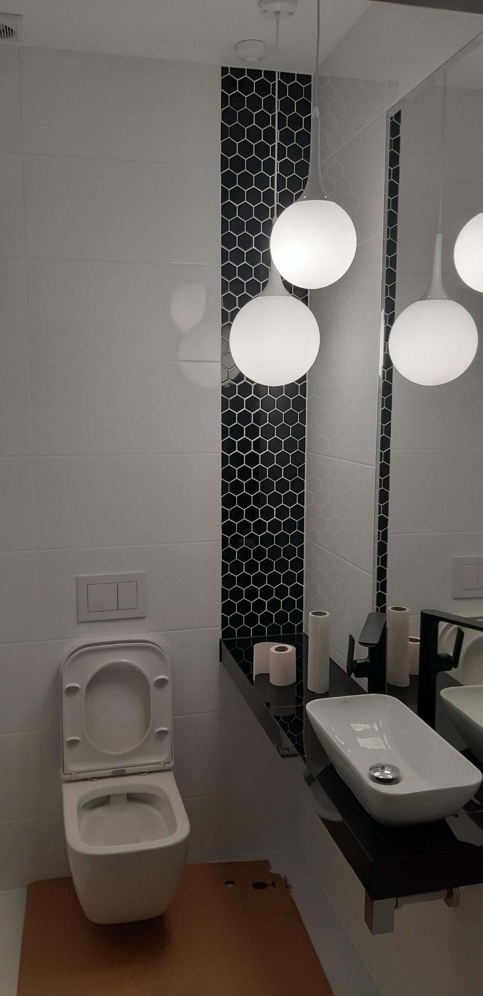 Nowoczesna łazienka z białymi płytkami, czarnym pasem dekoracyjnym w heksagony i wiszącymi lampami kulami, widoczna toaleta podwieszana z otwartą klapą i umywalka nablatowa z czarną armaturą.