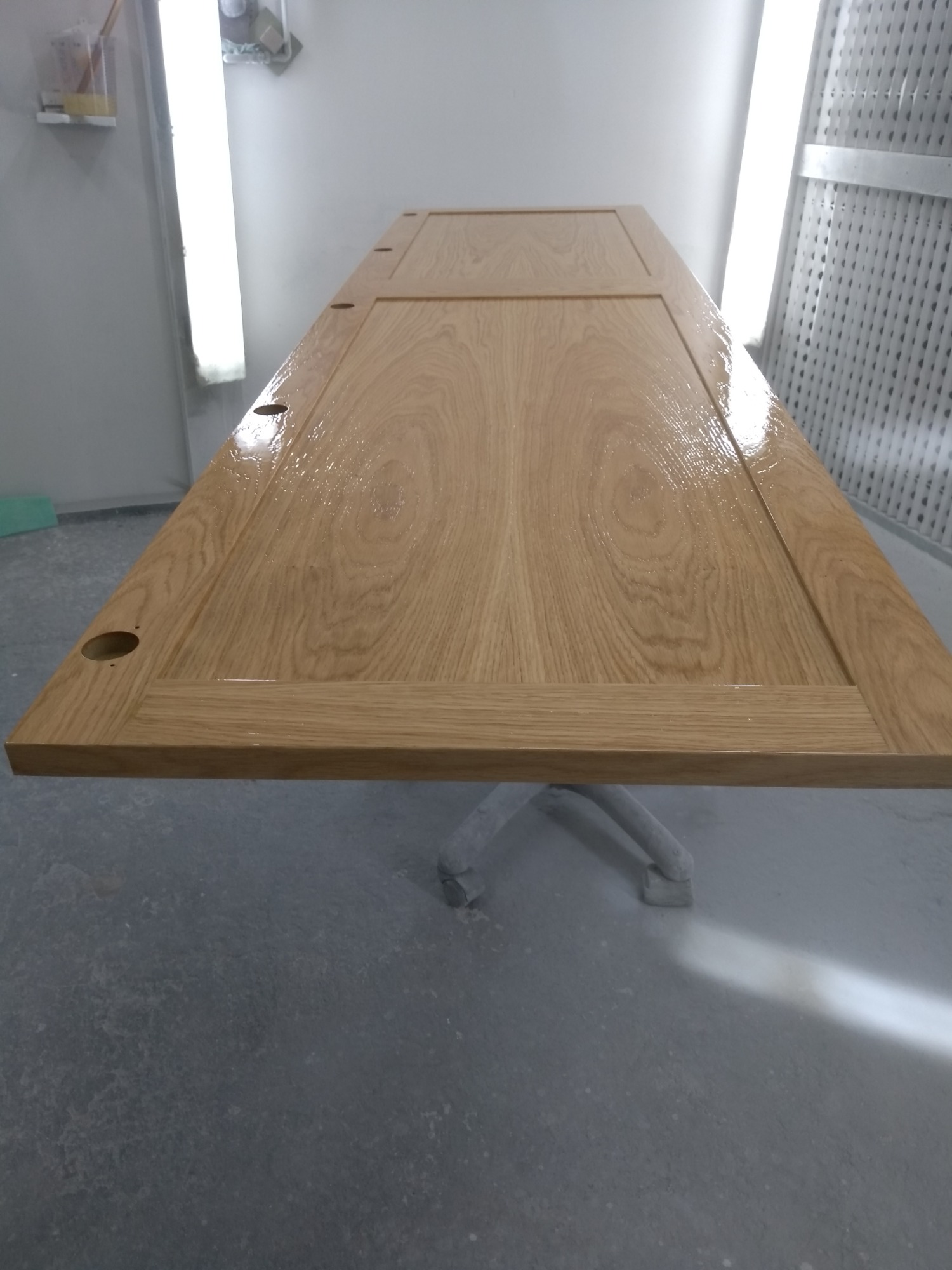 Lakierowane drzwi z jasnego drewna, leżące poziomo na metalowym stojaku w lakierni, widoczne otwory na klamkę i zamek.