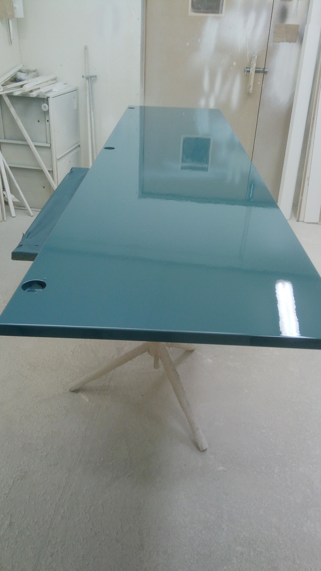 Turkusowy blat stołu z otworami, świeżo polakierowany, ustawiony na białym kozłach w warsztacie.