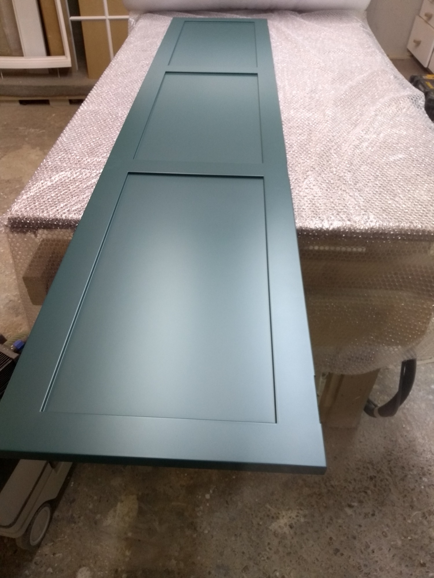 Pomalowane na ciemnozielono, gotowe do montażu, dwuczęściowe drzwi szafki z frezowanymi panelami, leżące na stole zabezpieczonym folią bąbelkową.