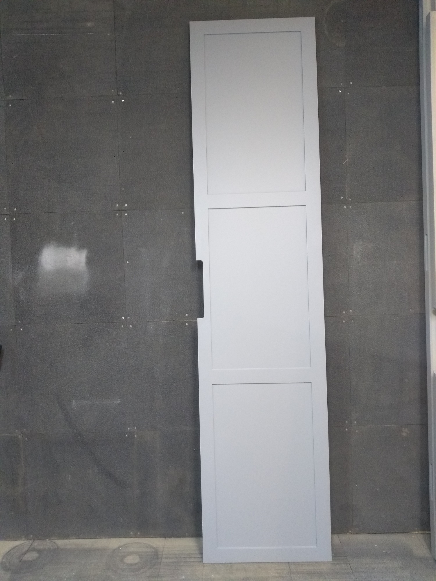 Świeżo pomalowane na szaro drzwi wewnętrzne z trzema panelami, opierające się o szarą ścianę z widocznymi punktami mocowania.