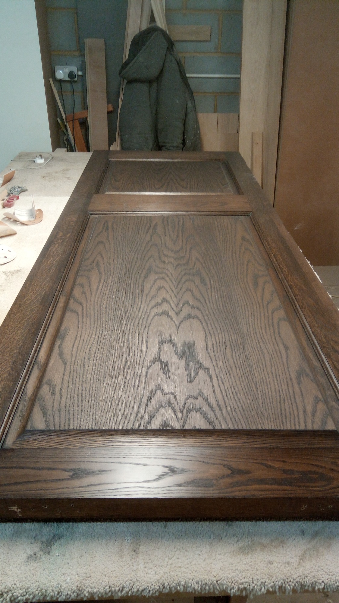 Odnowione drewniane drzwi z wyraźnym usłojeniem, leżące na stole w warsztacie, w tle narzędzia i ubranie robocze.