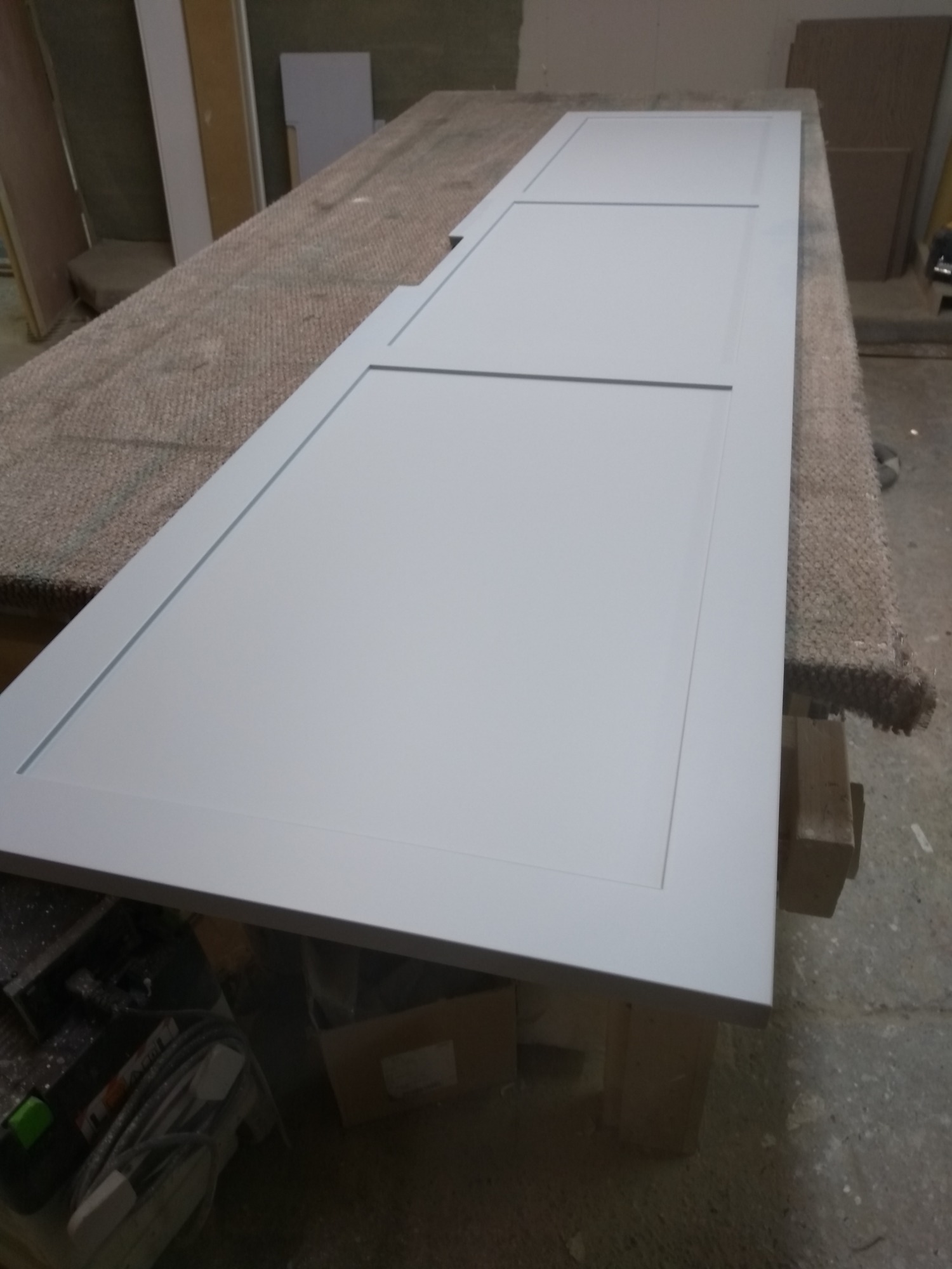 Pomalowane na szaro, segmentowe drzwi wewnętrzne, leżące na roboczym stole w warsztacie. Widoczne niedoskonałości w strukturze blatu stołu.