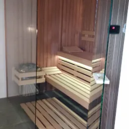 Wnętrze sauny z jasnego drewna z przeszklonymi ścianami, widoczne ławy, podświetlenie LED i kamienie na półce.